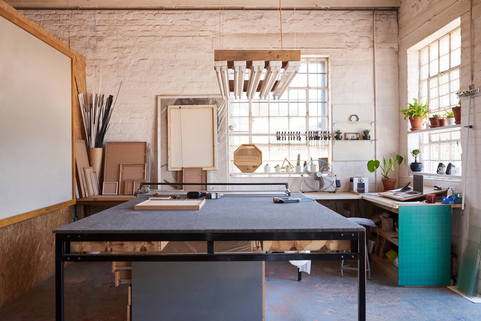 Atelier d'artisan avec établi noir central, murs en briques peintes blanches, grandes fenêtres industrielles
