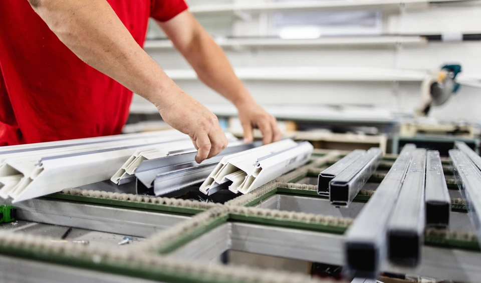 Atelier de fabrication industrielle avec ouvrier en t-shirt rouge assemblant des profilés aluminium sur établi