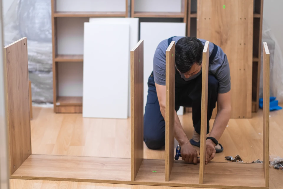 Un homme assemble des meubles en bois dans un atelier intérieur, agenouillé sur un parquet, utilisant un tournevis électrique sur une structure de meuble modulaire